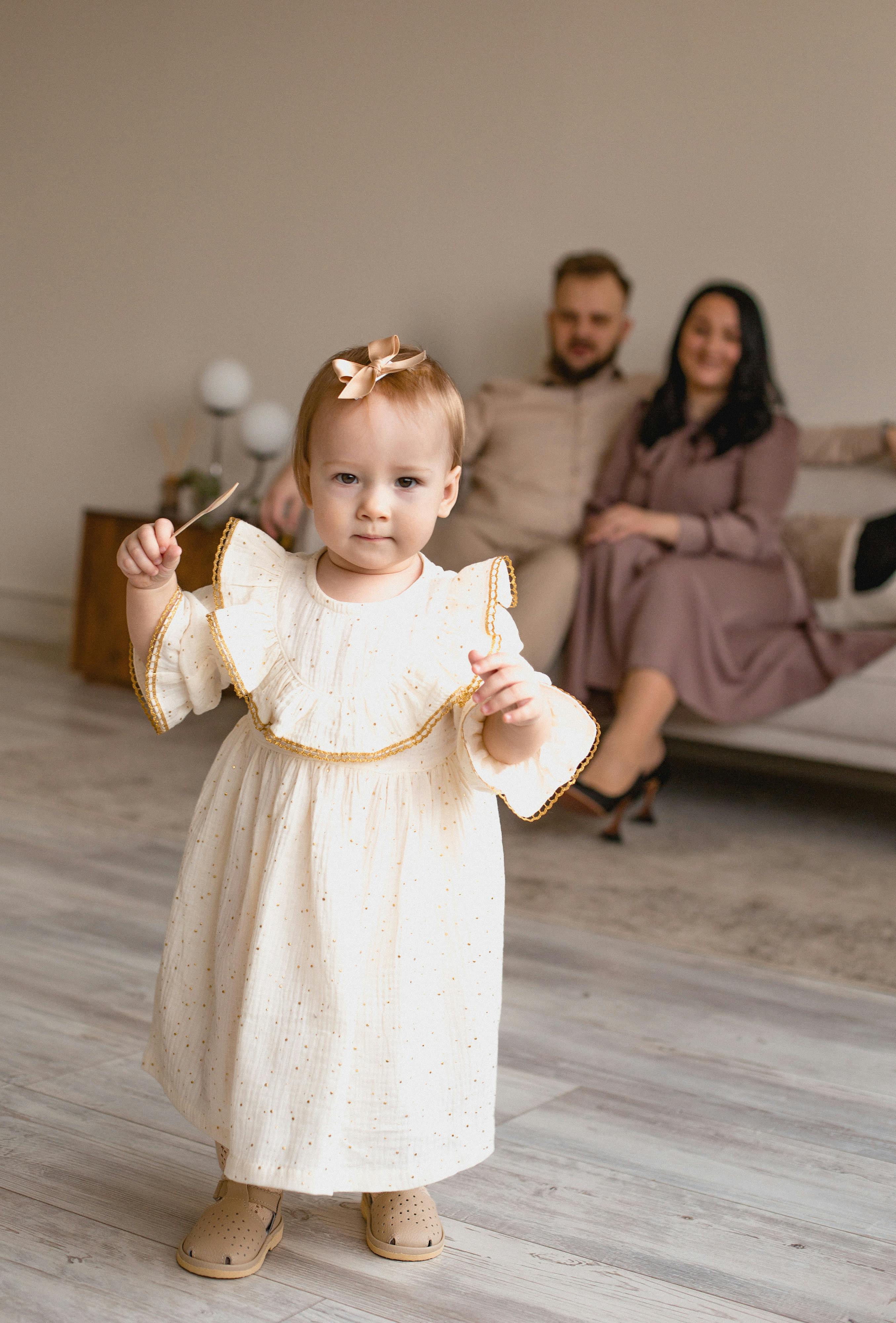 Adorable toddler in a dress standing indoors with parents in the background, exuding warmth and family togetherness.